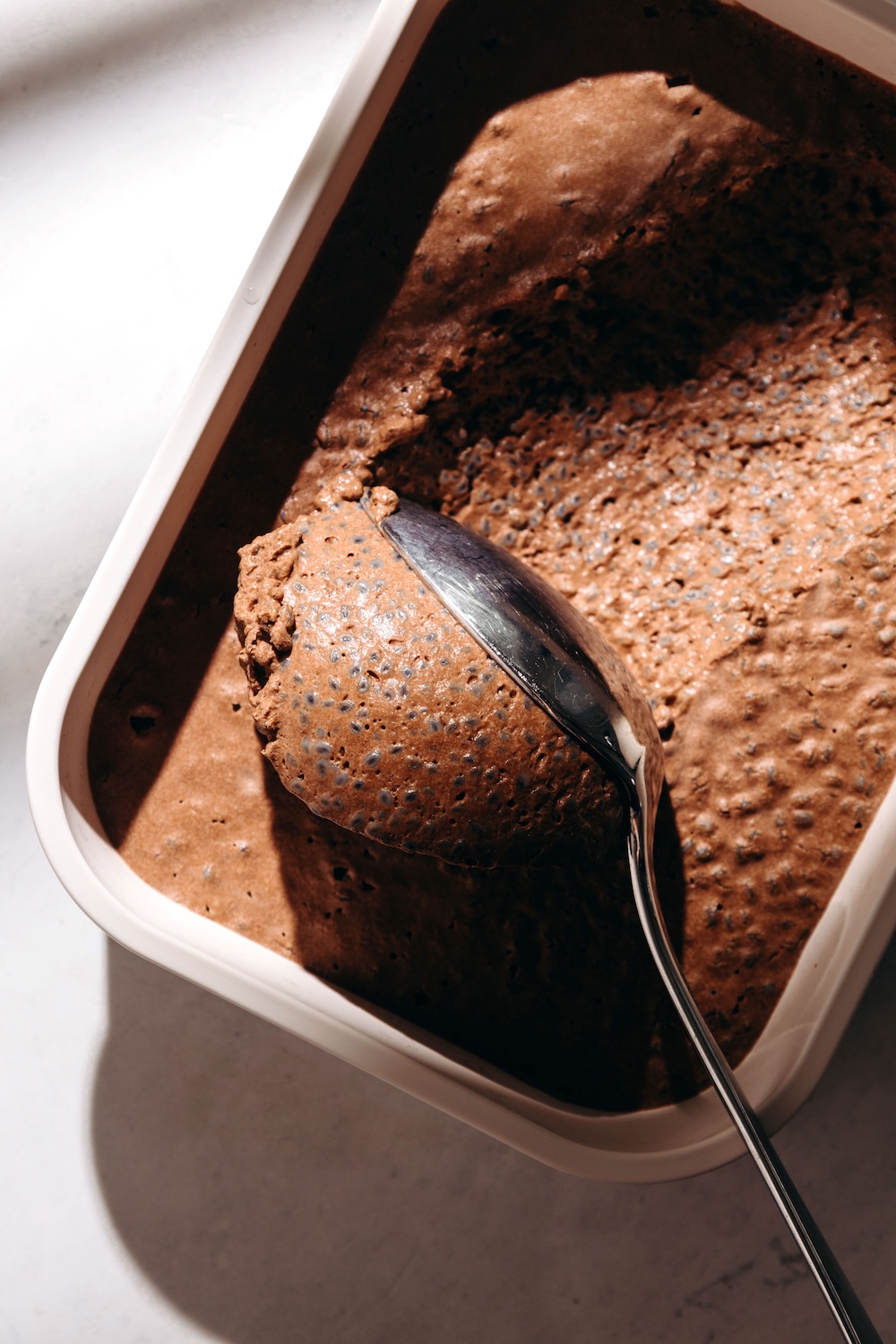 Chocolate basil seed pudding in a white container being scooped with a spoon, showing a thick, creamy texture with visible basil seeds throughout.
