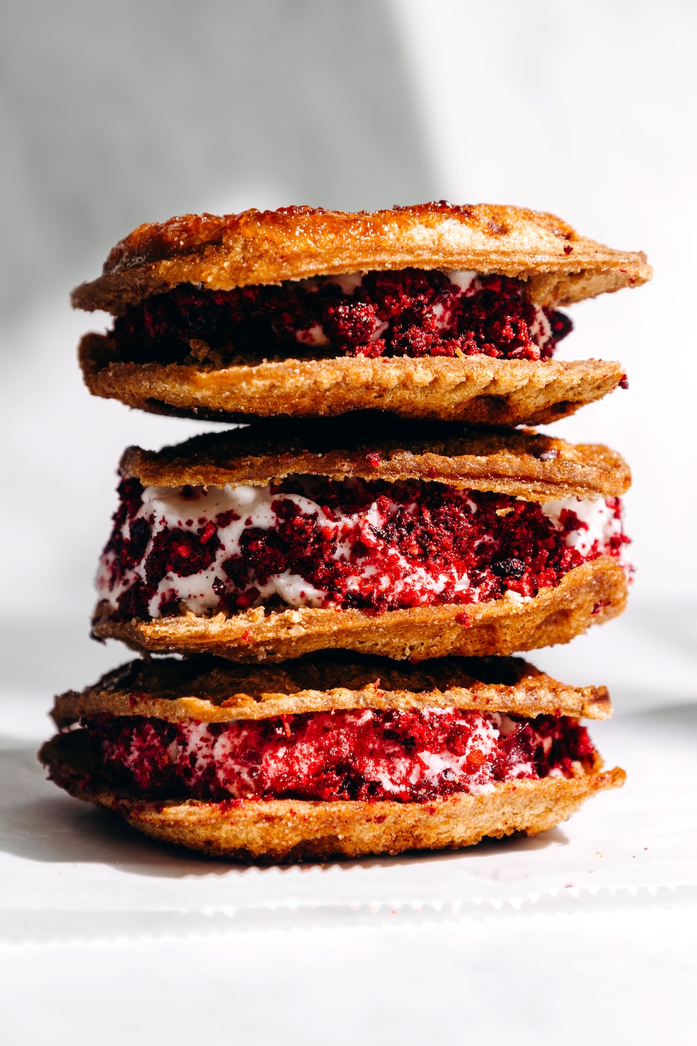 Stack of three waffle iron PB&J ice cream sandwiches made with SunButter Jammies, filled with dairy-free vanilla ice cream and coated in freeze-dried fruit dust on a white surface