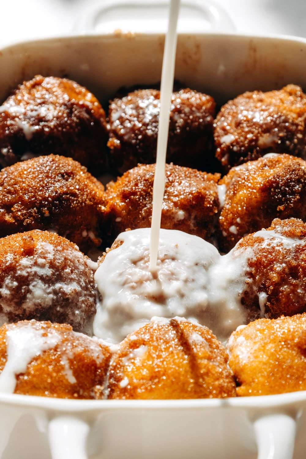 Coconut milk glaze being poured over pumpkin cinnamon roll bites in a white baking dish.