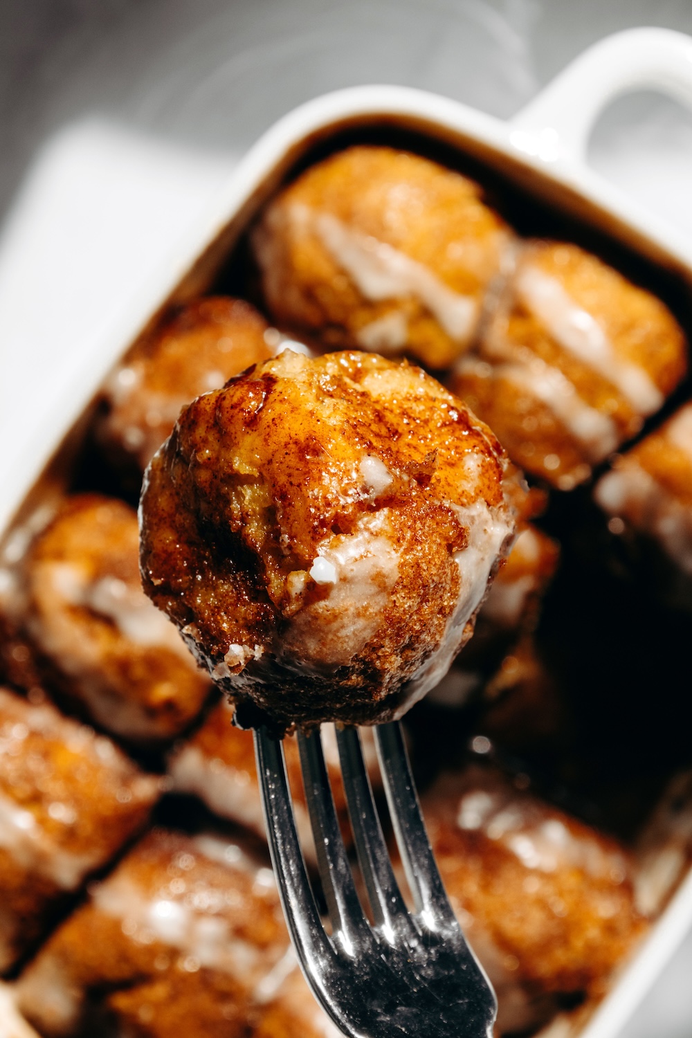 A glazed pumpkin cinnamon roll bite on a fork above a baking dish of more bites.