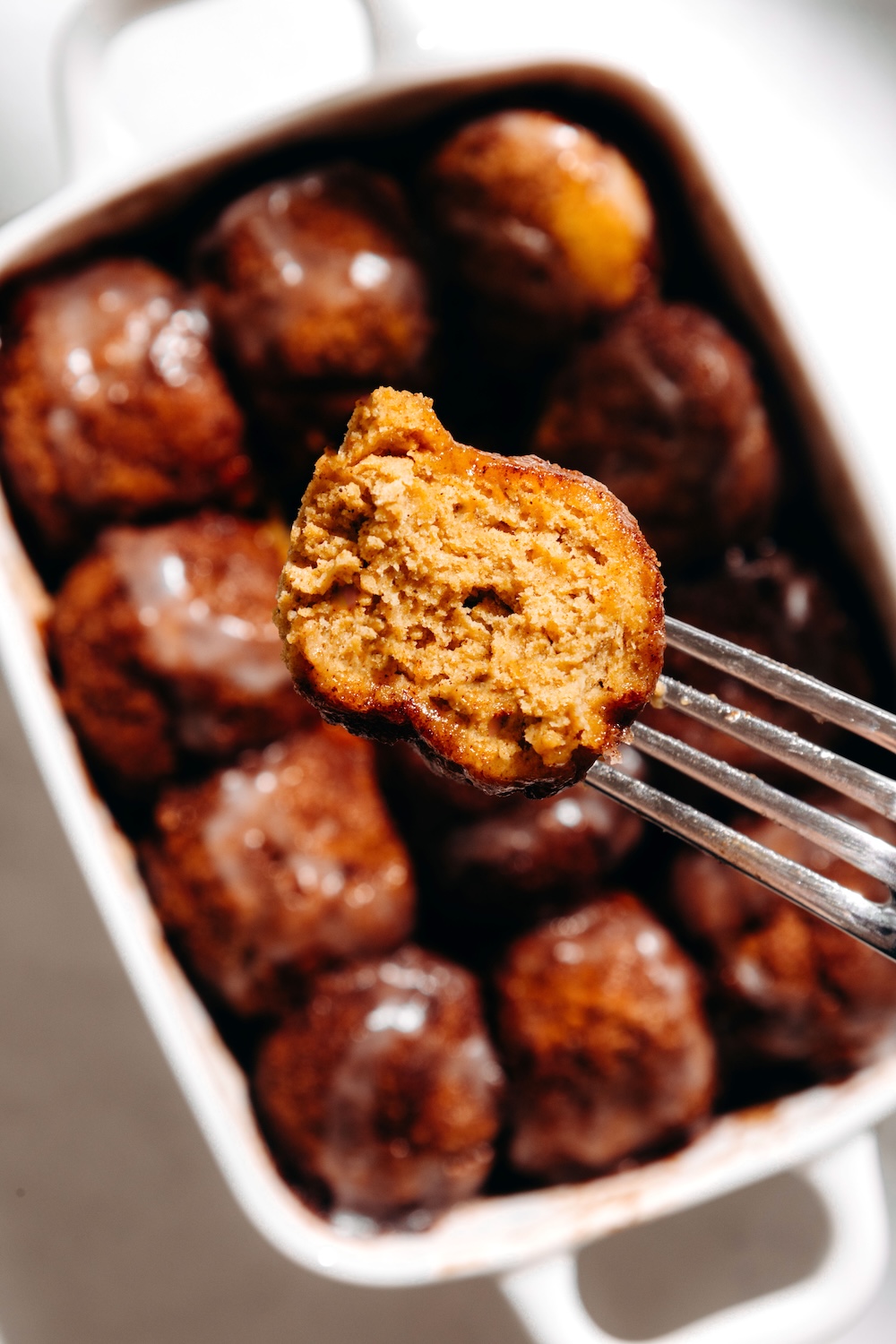 A pumpkin cinnamon roll bite on a fork held over a dish of glazed gluten-free cinnamon roll bites.