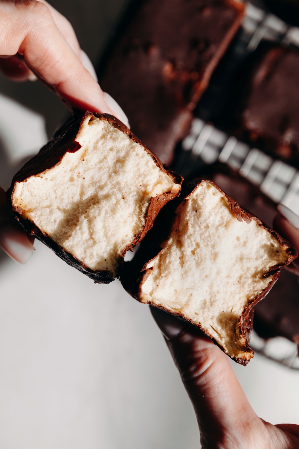 Hands breaking a chocolate-dipped protein marshmallow in half to show its fluffy, airy interior.