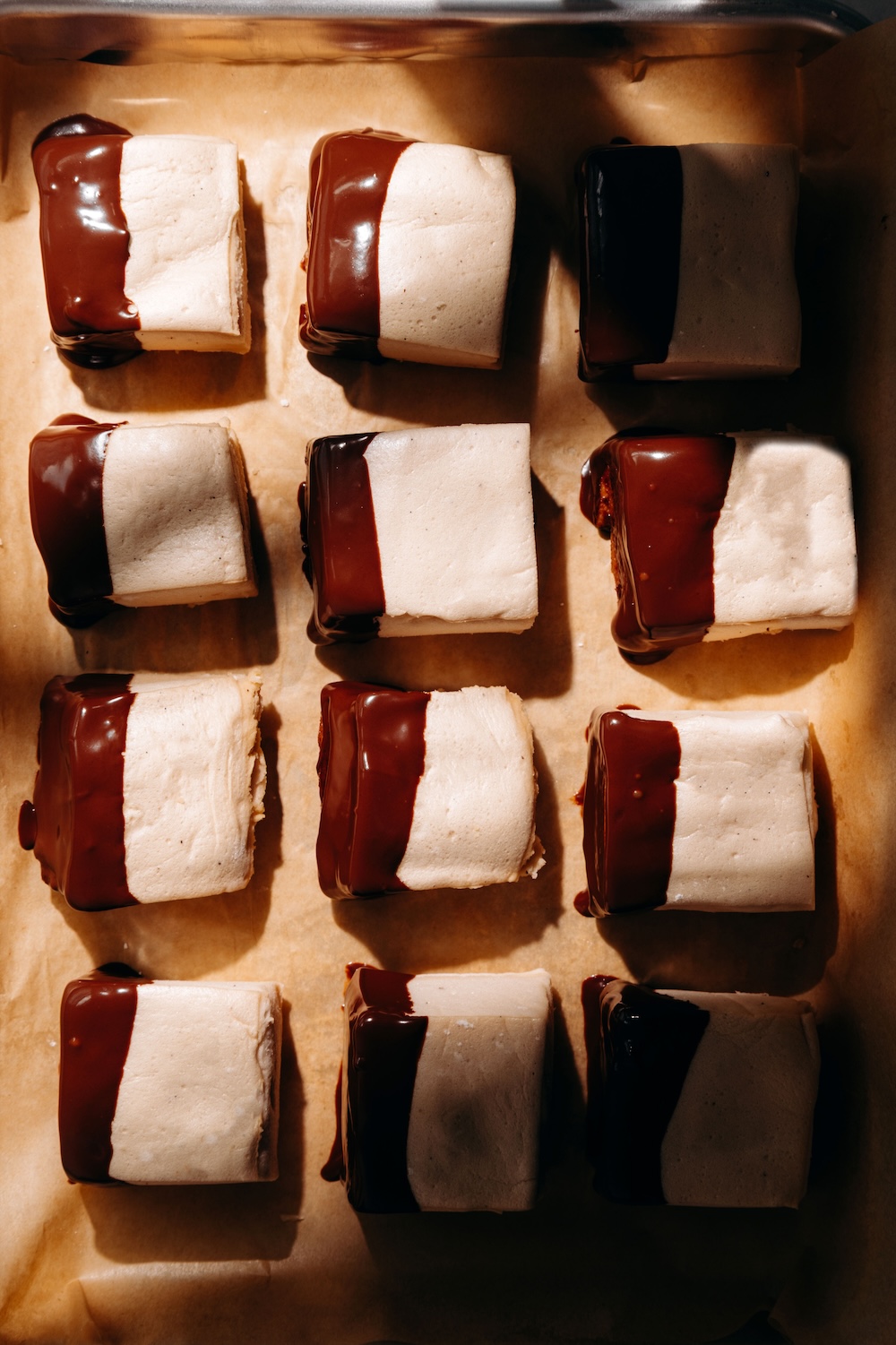 Protein marshmallows half-dipped in melted chocolate, lined up on a parchment-lined baking sheet.